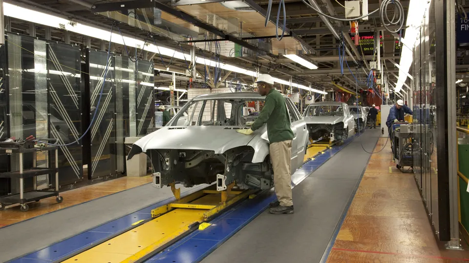 Car bodies moving down an assembly line in a vehicle plant, a worker in a hard hat at the first one