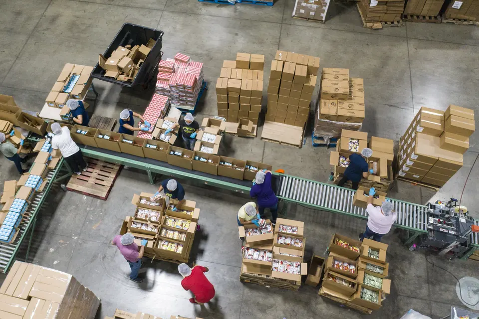 Overhead view of a warehouse floor: staff packing orders along roller conveyors among pallets of cartons